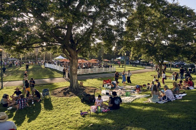 Visitors on blankets on green grass, surrounded by trees, and a fenced area with umbrellas in the distance