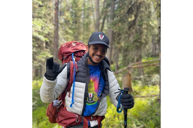 Aditya Srinath wearing a baseball hat, sunglasses, and a backpack in the woods