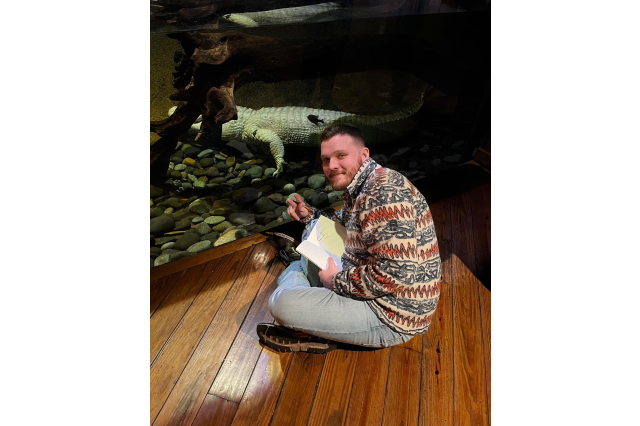 Cameron Clow sitting cross-legged on a wood floor in front of an alligator behind glass