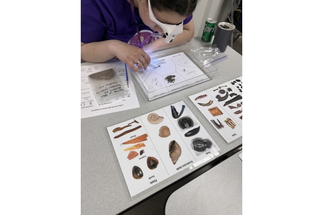Student wearing a headlamp looking over a tray of micro fossils with photos, of fossils on a white background , in front of the tray