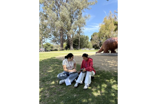 Two students sitting on the grass with one holding a clipboard and paper and the other holding a fossil skull