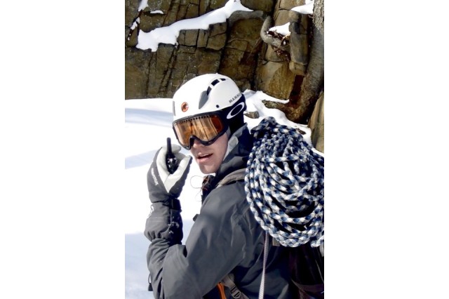 Matt Davis wearing a helmet and goggles and holding a walkie talkie, looking back towards in the camera in a snowy, rocky landscape