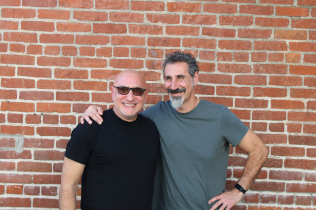 Kavat founders Serj Tankian and Ara Ajounian stand in their back patio in front of a brick wall.