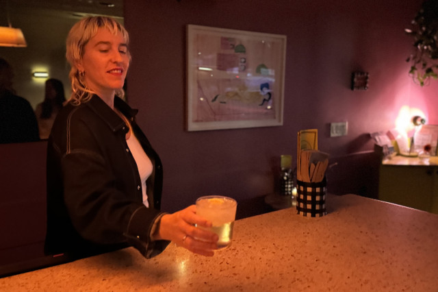 Blond woman in a black long sleeve shirt, standing behind a wooden bar, holding a cocktail glass.