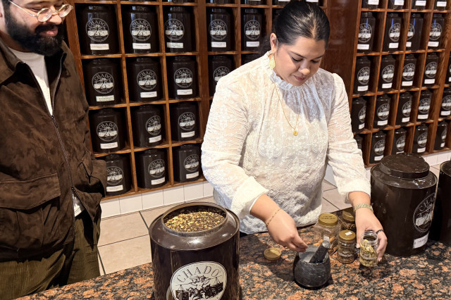 Woman standing at a marble table, combining tea leaves in a mortar and pestle, while a man stands next to her, smiling down at her.