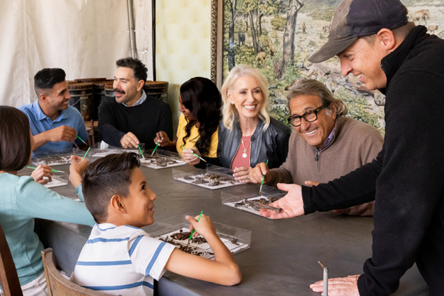 Excavator wearing a baseball cap and extending hand towards a group of visitors sorting fossils at a table