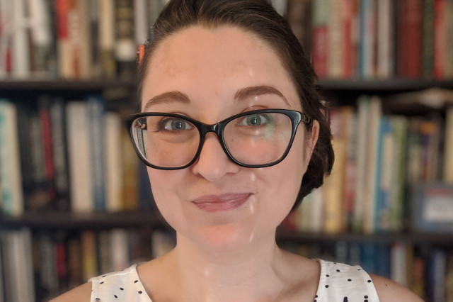 Kriss Leftwich wearing black eyeglasses and a black and white polka dot tank top with shelves of books in the background