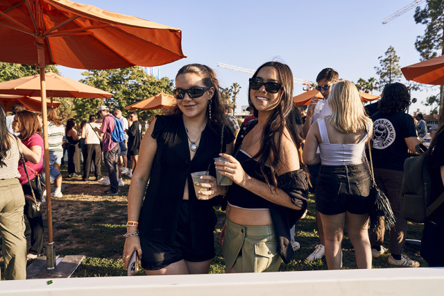 Two women holding drinks with their arms around each other, with orange umbrellas and other visitors in the background
