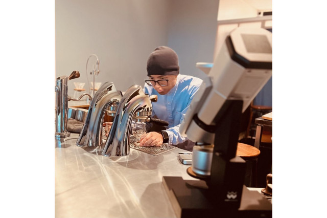 Man in glasses and a beanie leaning over a coffee bar making an artisanal cup of coffee.