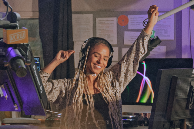 DJ Novena Carmel with arms raised, surrounded DJ equipment and computer