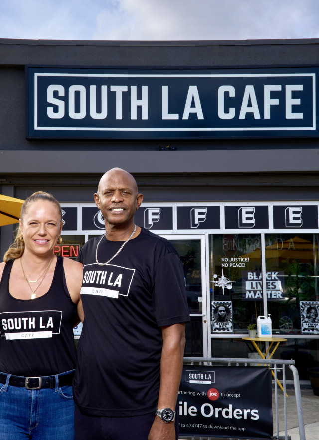 Man and woman standing in front of a grey-colored corner coffee shop with the sign "South LA Cafe" above the door. They are both wearing t-shirts with the cafe logo on them; the woman is holding a coffee cup.