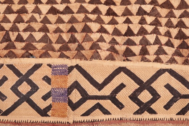 Pattern of small brown triangles above strip of x outline patterns. Section of a Kuba wrap skirt from the Kasai Province of the Democratic Republic of the Congo (DRC), c. 1900. It is made of stitched barkcloth, woven raffia, and cut pile embroidered borders (the cut pile stitching resembles velvet).