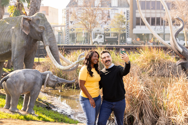 Couple holding up a camera to take a photo of themselves in front of mammoth sculptures
