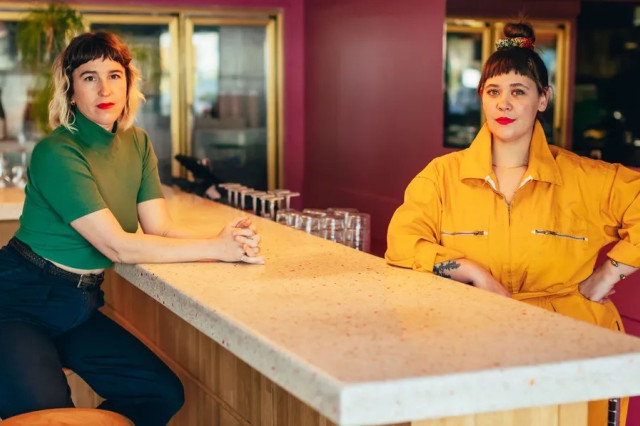Two women on either side of a marble bar table, one sitting, the other standing. 