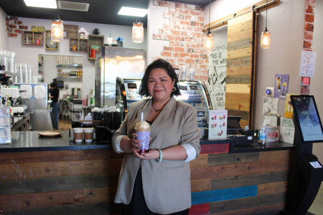 Filipino woman with short hair and a tan blazer holding a purple halo-halo drink in front of a cafe counter.