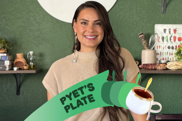 Woman with long, dark hair over one shoulder, standing in front of a green wall with shelves of cups, cookbooks, and plants. 