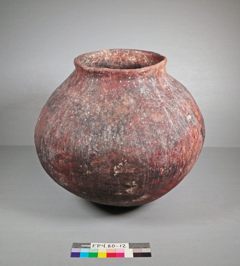 The object photographed is a globular ceramic jar or olla with a wide, rounded body and a short, flared neck. It sits directly on its curved base without a stand. The vessel has a worn, weathered appearance with a complex, mottled surface of deep earthy tones.