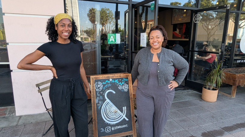 Two black women stand next to a black and brown A-frame sign with an ampersand and floral designs in front of the "Sip & Sonder" coffee shop.