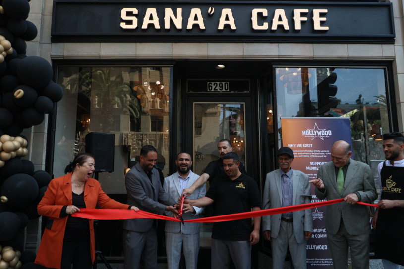 A group of people stand outside the storefront of Sana'a Cafe, holding large scissors and cutting a wide red ribbon to celebrate the grand opening of the Hollywood location.