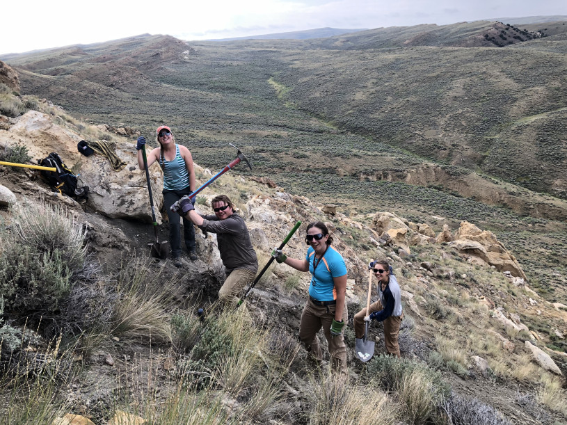 A line of five people holding digging tools walk on a rocky mountain landscape