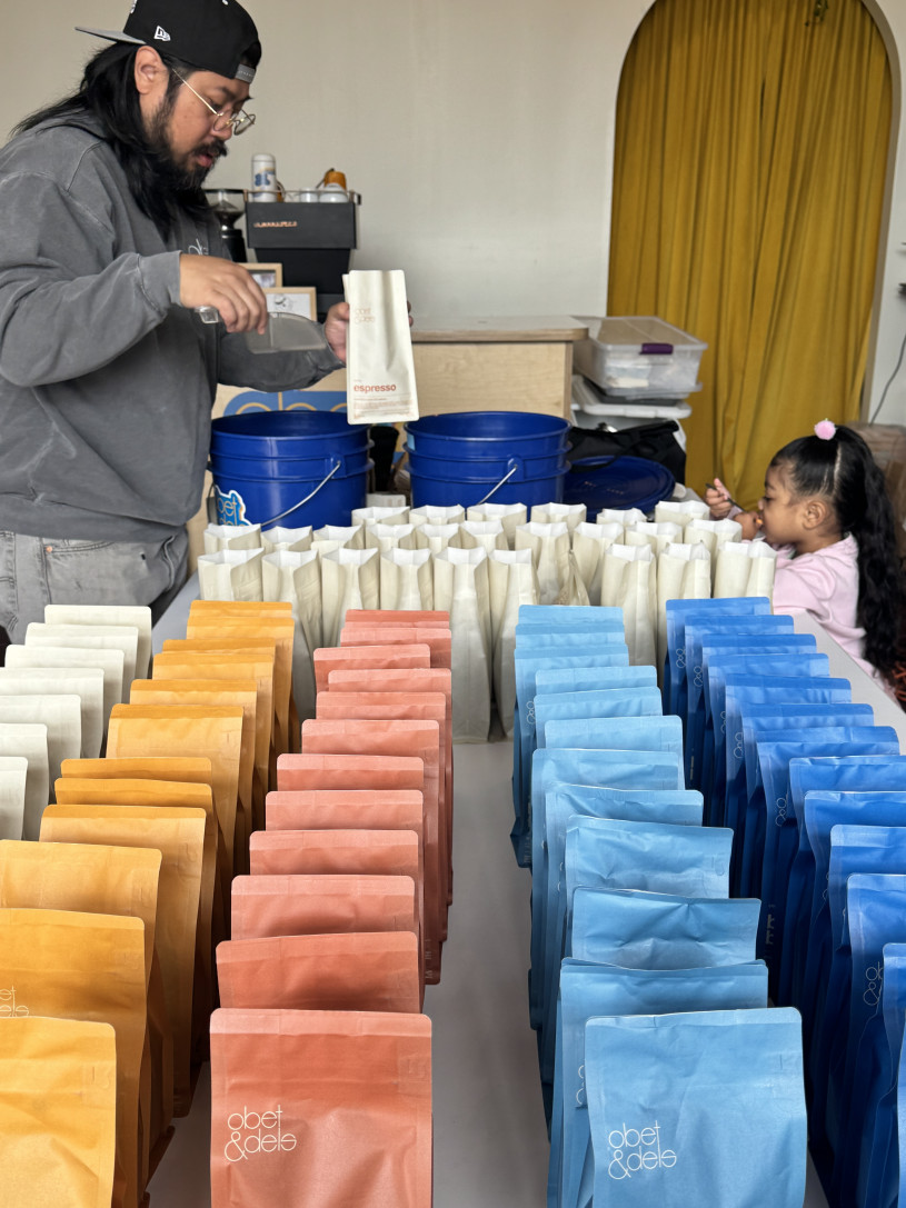 A mand and small child stand behind a table of rows of brightly colored coffee bean bags. 